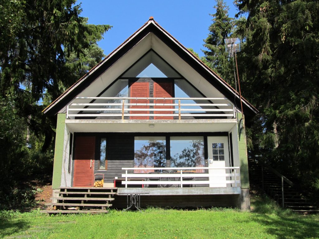 Kaksikerroksinen harjakattoinen rinteeseen rakennettu lomamökki suurten kuusten välissä. Etualalla nurmikkoa joka viettää kohti katsojaa.